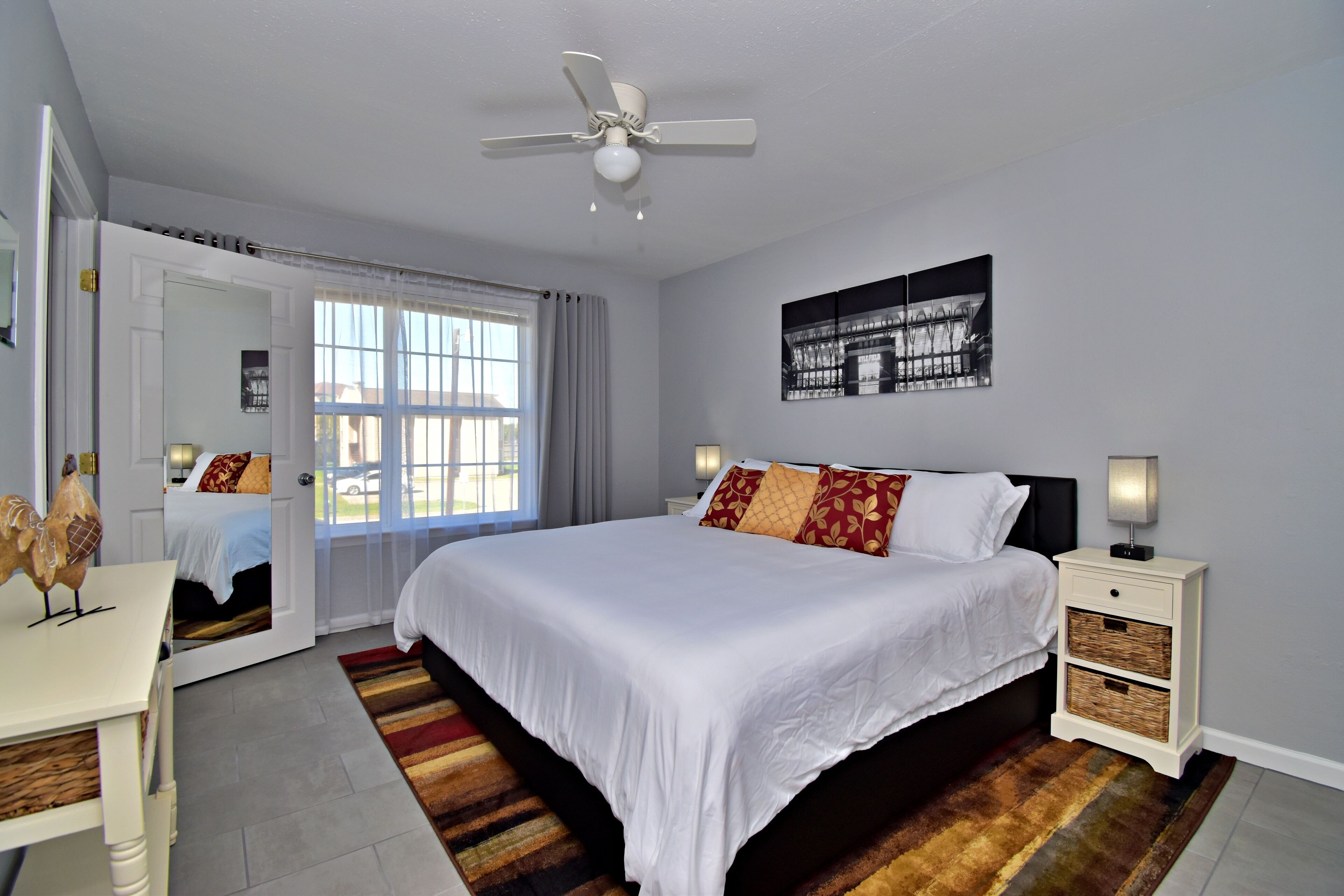 Une chambre à coucher comprenant un lit, des tables de chevet, un ventilateur de plafond, une fenêtre avec des rideaux, un tableau accroché au mur et une porte avec un miroir.