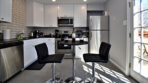 A modern kitchen with white cabinets, stainless steel appliances, a black countertop, and two bar stools. - ✪ King Beds ✔ 2 Bdr Townhouse w/ Private Backyard (Bryan)