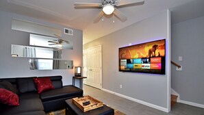 A modern living room with a flat-screen TV mounted on the wall, a ceiling fan, a black sectional sofa, and a wooden coffee table. - ✪ King Beds ✔ 2 Bdr Townhouse w/ Private Backyard (Bryan)