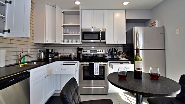 A modern kitchen with white cabinets, stainless steel appliances, a built-in oven, and a dining area with a round table and chairs. - ✪ King Beds ✔ 2 Bdr Townhouse w/ Private Backyard (Bryan)