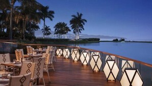 Eine Terrasse mit Tischen und Stühlen, Palmen und Blick auf Wasser und Berge bei Nacht.