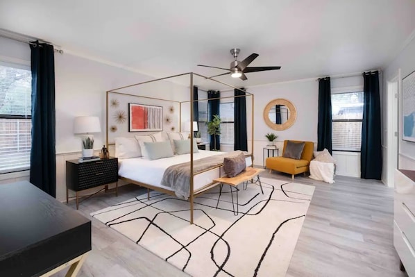 A modern bedroom with a four-poster bed, a ceiling fan, a side table with a lamp, a small sofa, and a round mirror.
