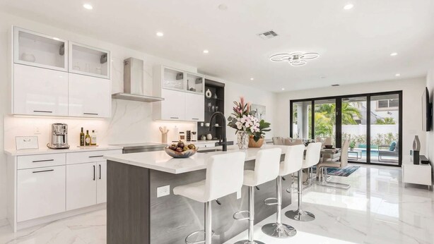 A modern kitchen with white cabinetry, a central island with bar stools, and a dining area with a view of the outdoors. - Luxury Paradise, walk to Las Olas -5 mins to Beach (Fort Lauderdale)