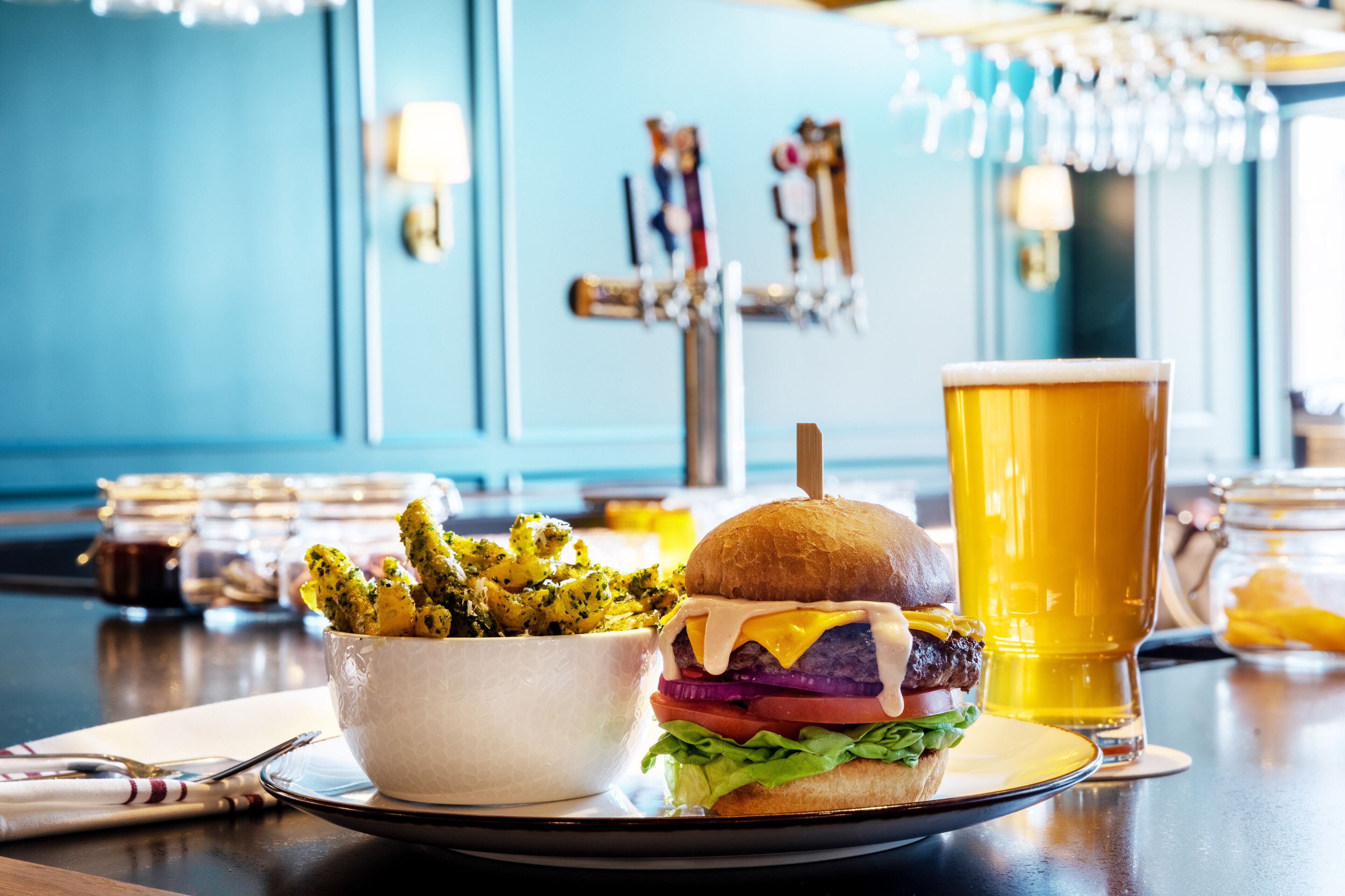 Un hamburger composé d’un pain, de laitue, de fromage et d’une galette, accompagné d’un verre de bière et d’un bol de frites, le tout posé sur une table.