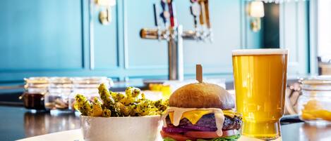 Un hamburger composé d’un pain, de laitue, de fromage et d’une galette, accompagné d’un verre de bière et d’un bol de frites, le tout posé sur une table.