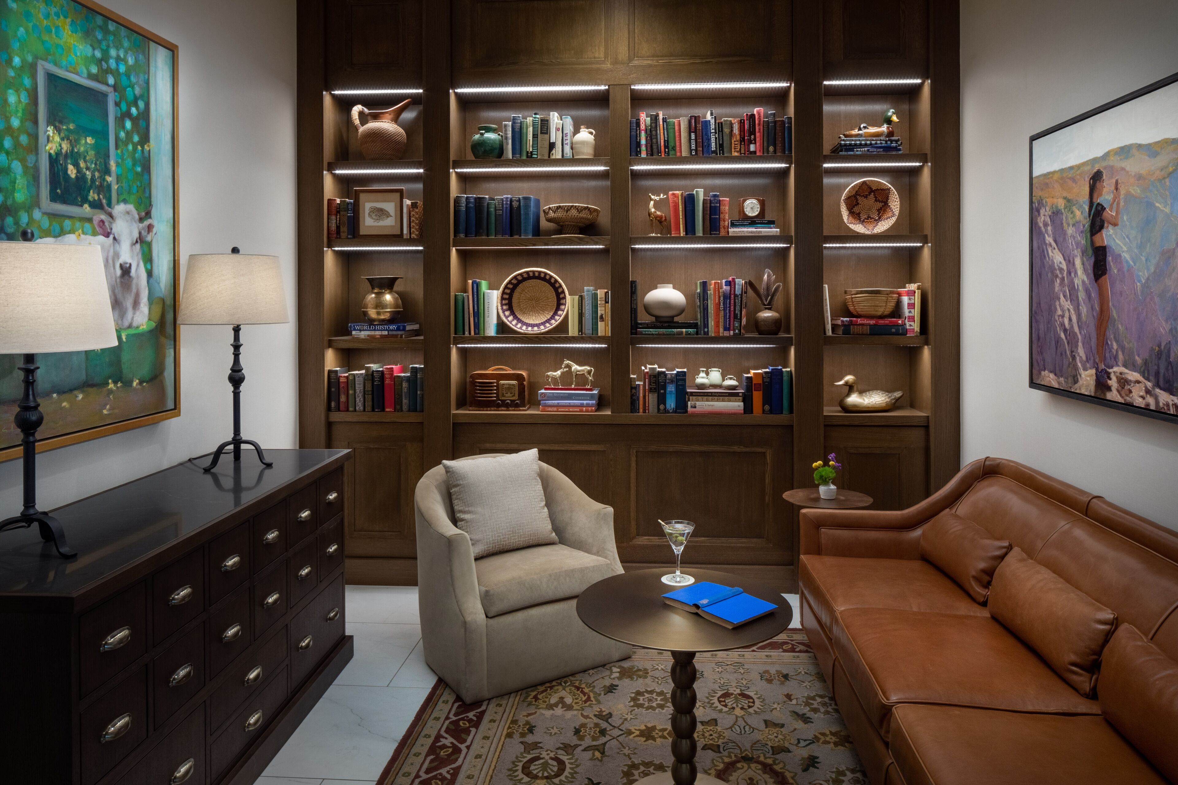 Un salon avec un canapé en cuir marron, un fauteuil beige, une bibliothèque en bois remplie de livres et d’objets décoratifs, une petite table d ’appoint en bois foncé sur laquelle se trouvent un livre bleu et un verre à martini, et un grand tableau accroché au mur.