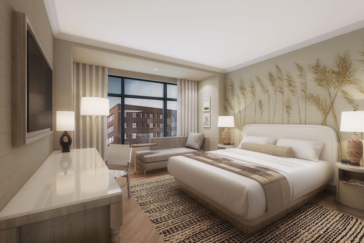 Una habitación de hotel moderna con una cama grande, dos mesitas de noche con lámparas, un escritorio, una silla y vistas a edificios por la ventana.