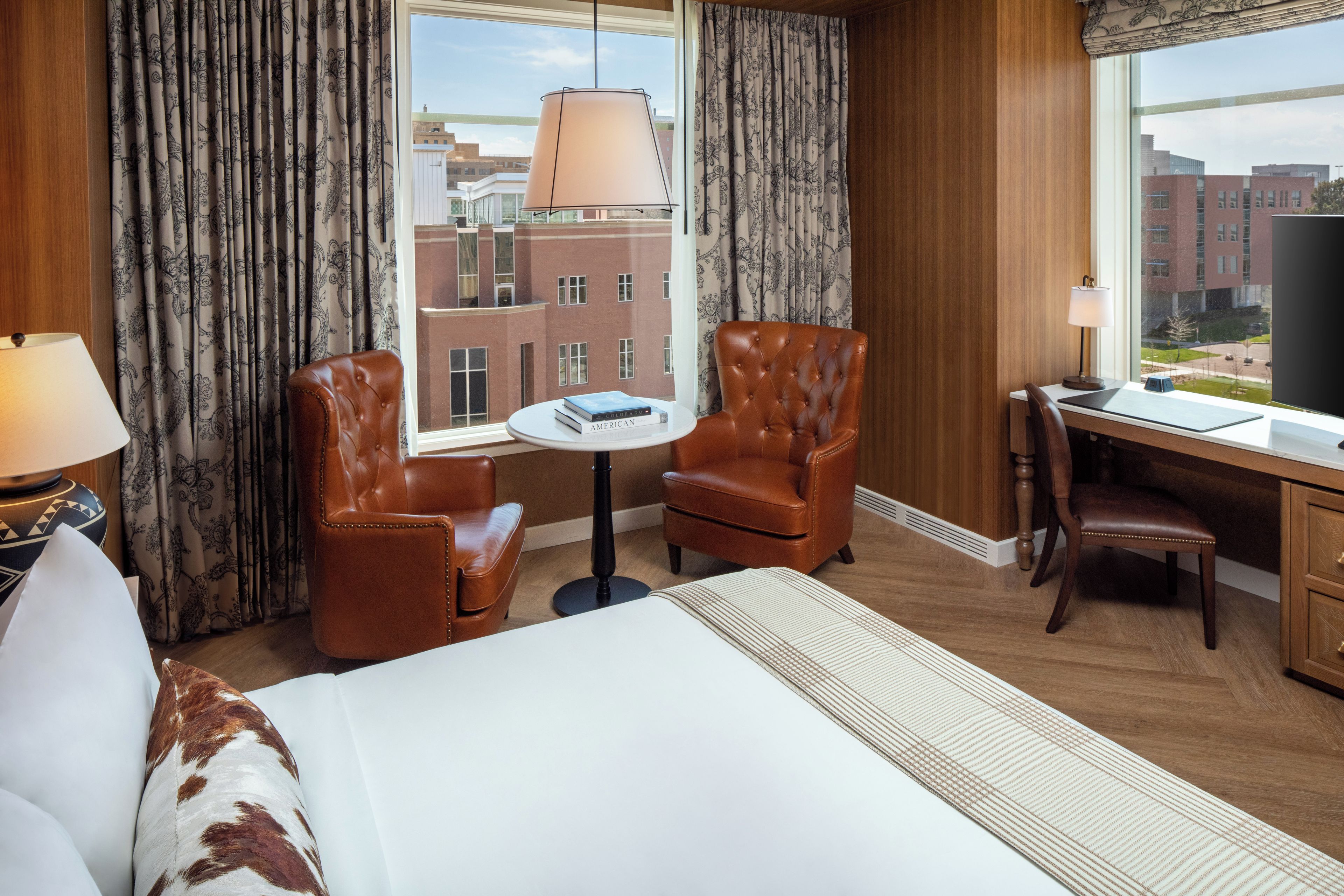Una habitación de hotel con una cama, dos sillones de cuero, una mesita, un escritorio y un ventanal con vistas a edificios.