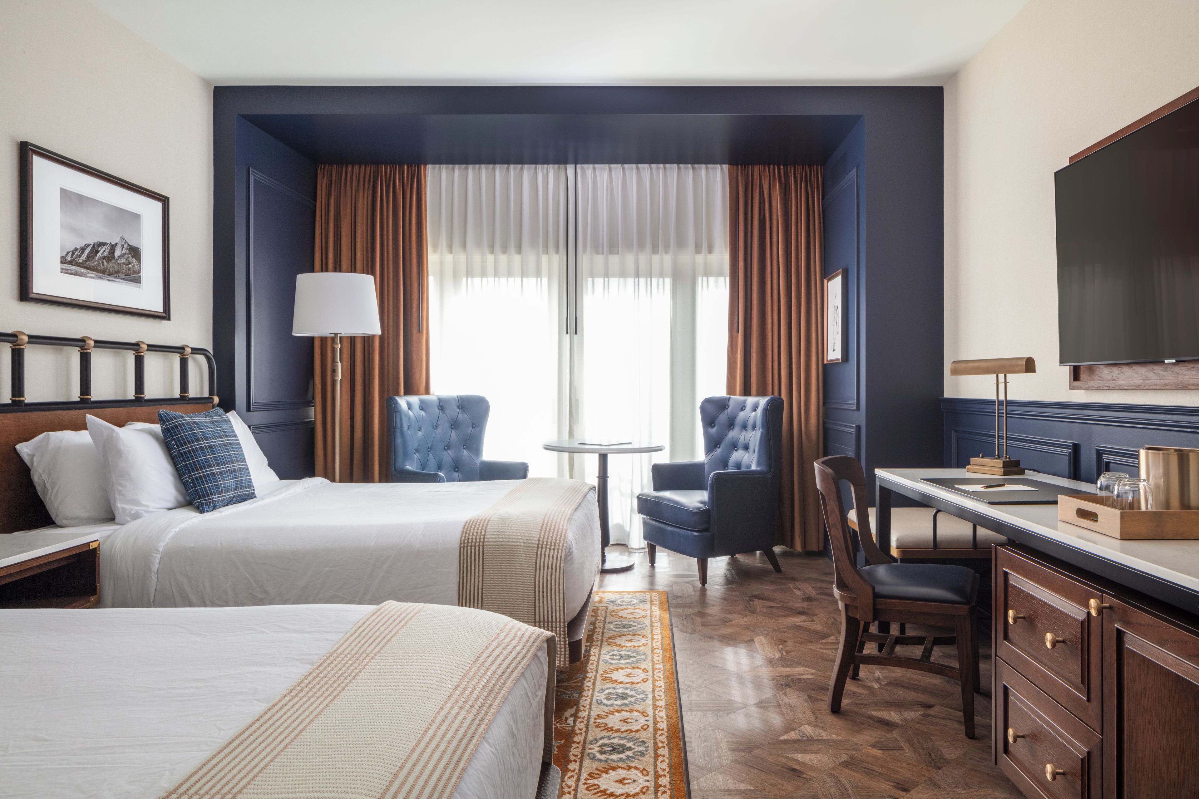 A hotel room with two beds, a desk, a chair, a television, and a window with curtains.