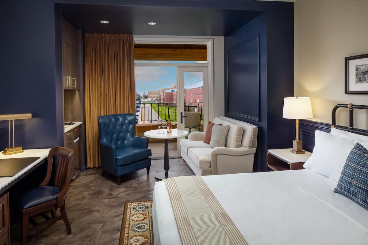 Una habitación de hotel con cama, un sillón azul, un sofá blanco, una mesita redonda y vistas a edificios a través de una ventana.