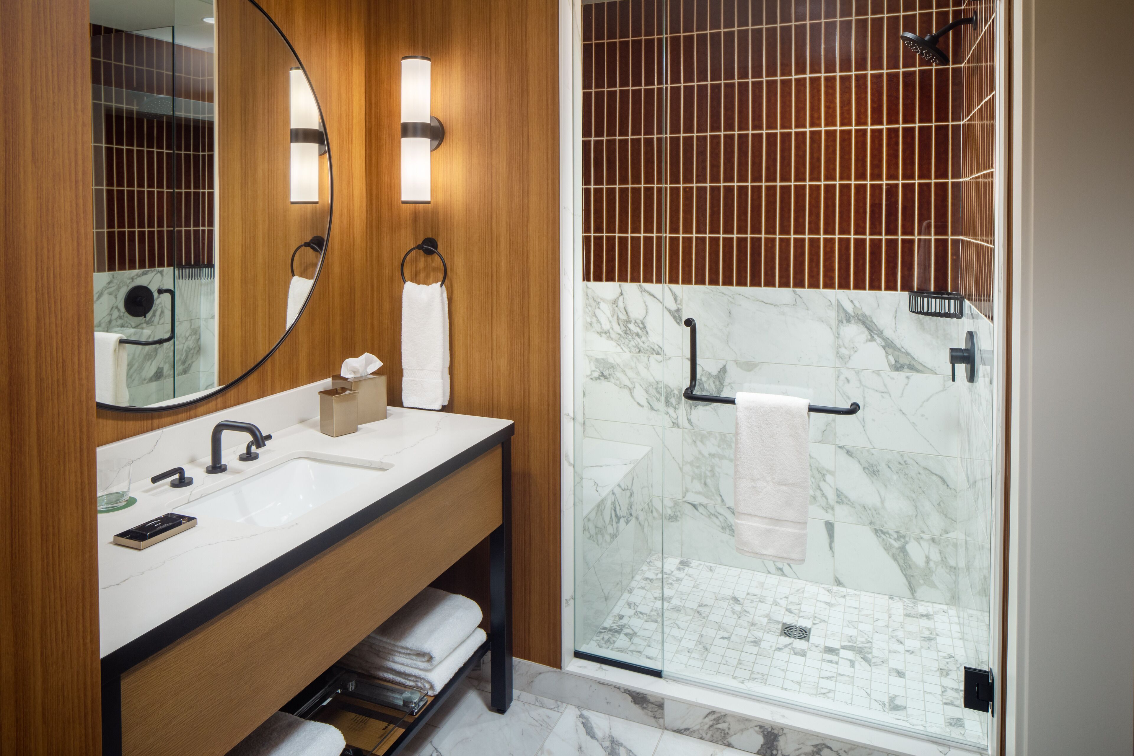 A modern bathroom with a wooden vanity, a large mirror, a glass shower enclosure, and marble tiles.