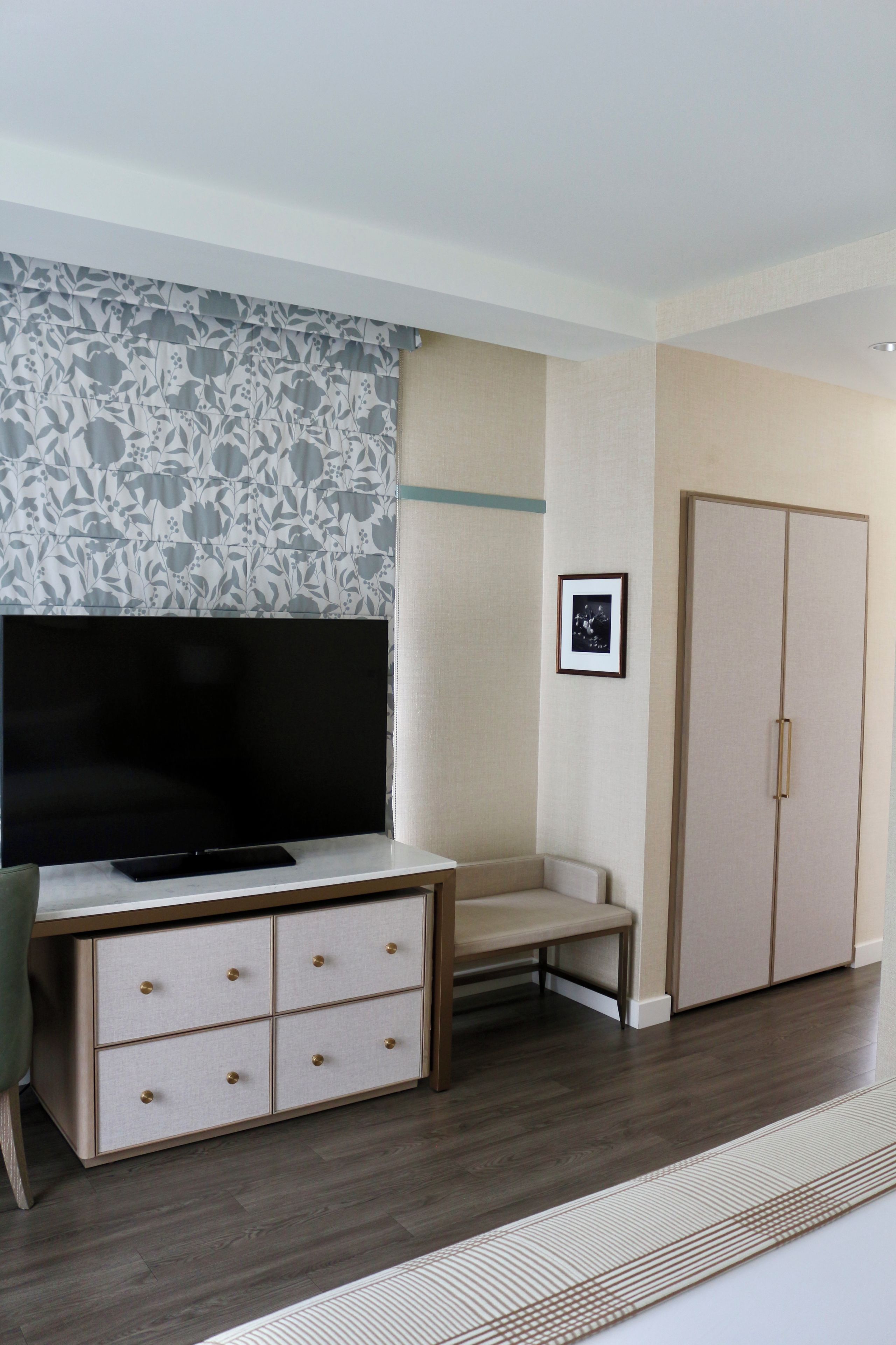 A room with a television on a stand, a desk with drawers, a chair, a wardrobe, and a framed picture on the wall.