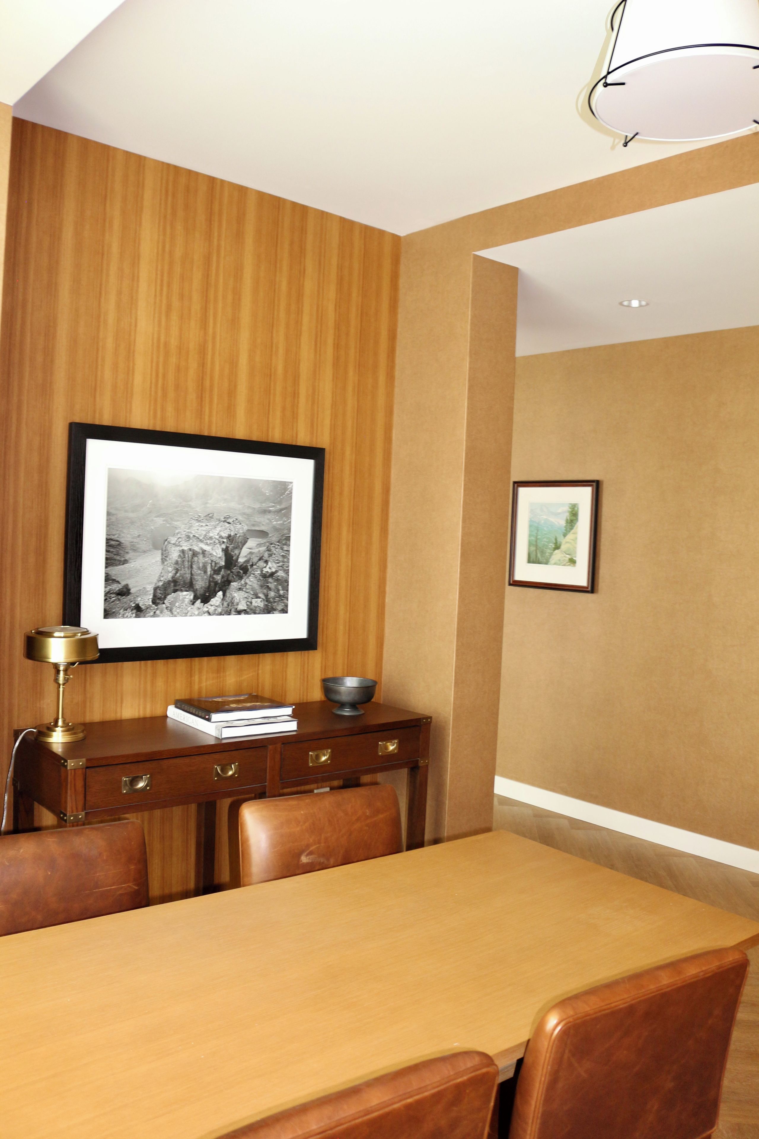A dining area with a wooden table, chairs, a framed photograph on the wall, and a desk with a lamp and books.
