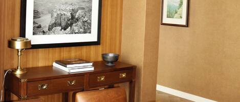A dining area with a wooden table, chairs, a framed photograph on the wall, and a desk with a lamp and books.