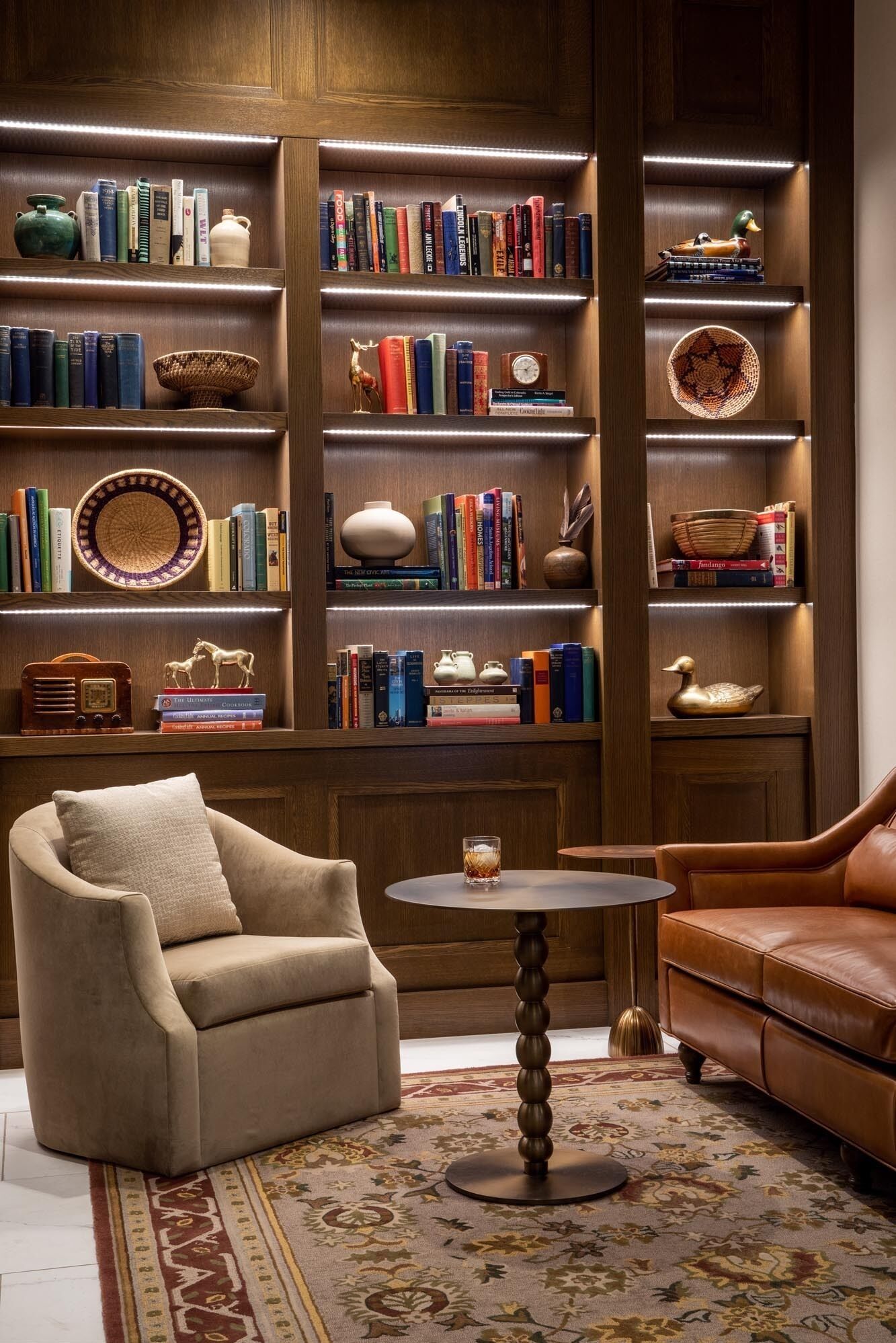 Un coin lecture confortable avec un fauteuil en cuir, une petite table ronde et une bibliothèque remplie de livres et d’objets décoratifs.