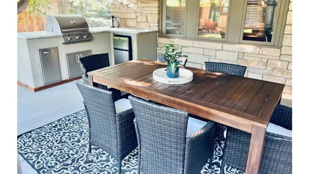 An outdoor dining area with a wooden table and wicker chairs, a grill, and a stone wall.