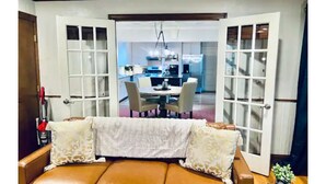 A living room with a brown leather sofa, a white throw blanket, and patterned pillows. There is a dining area visible through an open door in the background.