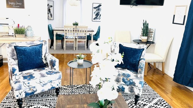 Un salon comprenant deux fauteuils à motifs, une table basse, un téléviseur, un coin repas avec une table et des chaises, et un téléviseur fixé au mur.