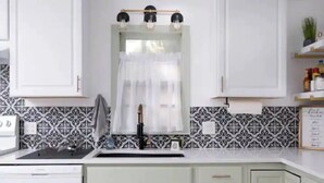 Une cuisine avec des meubles blancs, un pare-feu noir et blanc à motifs, une fenêtre avec un rideau blanc et une étagère contenant divers objets.