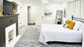 Une chambre à coucher avec un lit, un ventilateur de plafond, une télévision, une cheminée et un couloir menant à une autre pièce.
