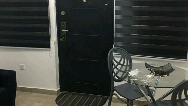 A room with a black door, a glass table with chairs, a black sofa, and a remote control on a black ottoman. - Ajay's Residence for short and long stays. (Tema)