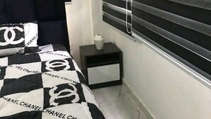A bedroom with a bed covered in a black and white checkered blanket, a nightstand with a vase, and a window with blinds. - Ajay's Residence for short and long stays. (Tema)