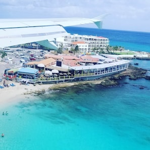 An aerial view of a coastal hotel with a large building, a beach area with umbrellas, and a clear view of the ocean.