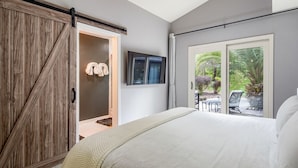 Un dormitorio con una cama grande, un armario con puerta corredera estilo granero, un televisor montado en la pared y un ventanal corredizo que da a un patio exterior con mobiliario.
