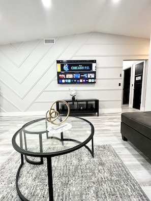 A modern living room with a glass coffee table, a flat-screen TV mounted on the wall, a black entertainment unit, and a grey sofa.