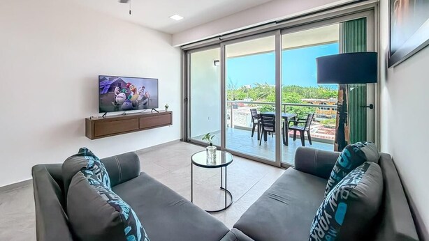 Ein modernes Wohnzimmer mit Sofa, Fernseher, Deckenventilator und einer Schiebeglas-Tür, die zu einer Terrasse im Freien führt.