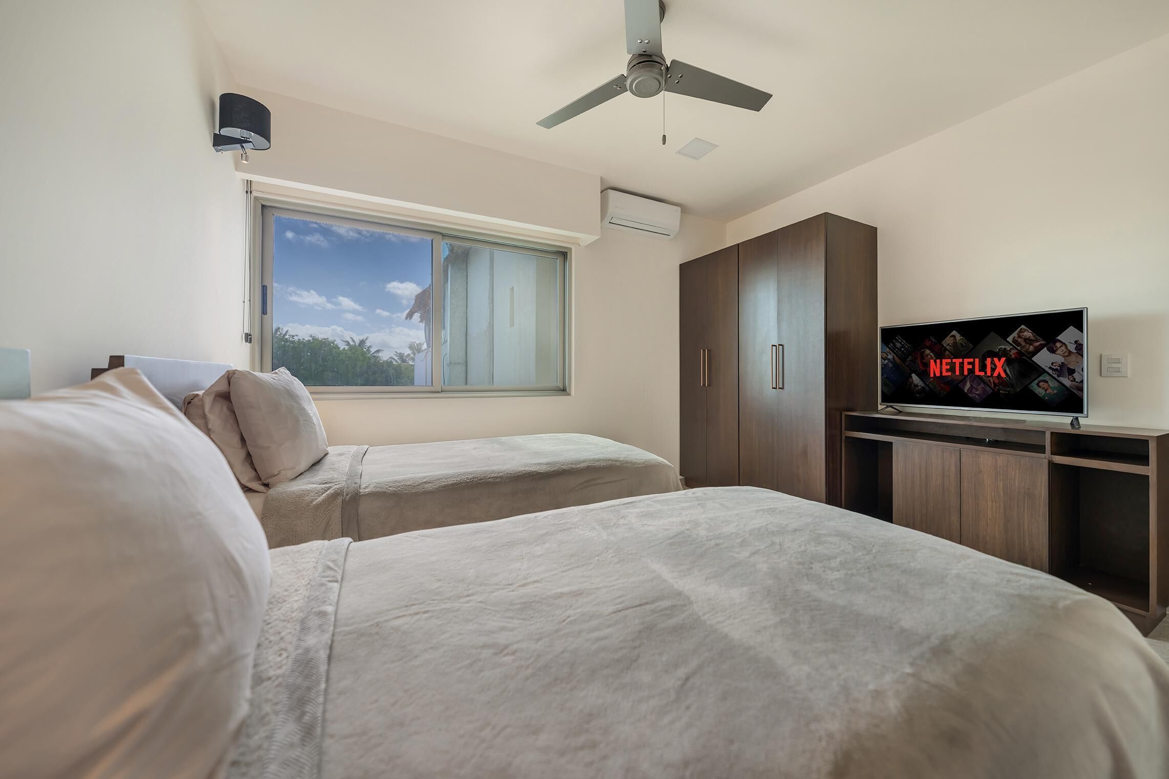 Habitació d'hotel amb dos llits, un ventilador de sostre, un televisor que mostra el logotip de Netflix i una finestra amb vista a l'exterior.