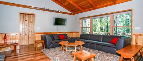 A spacious living room with a wooden ceiling, a large window, a black leather sofa, a wooden coffee table, and a television mounted on the wall.