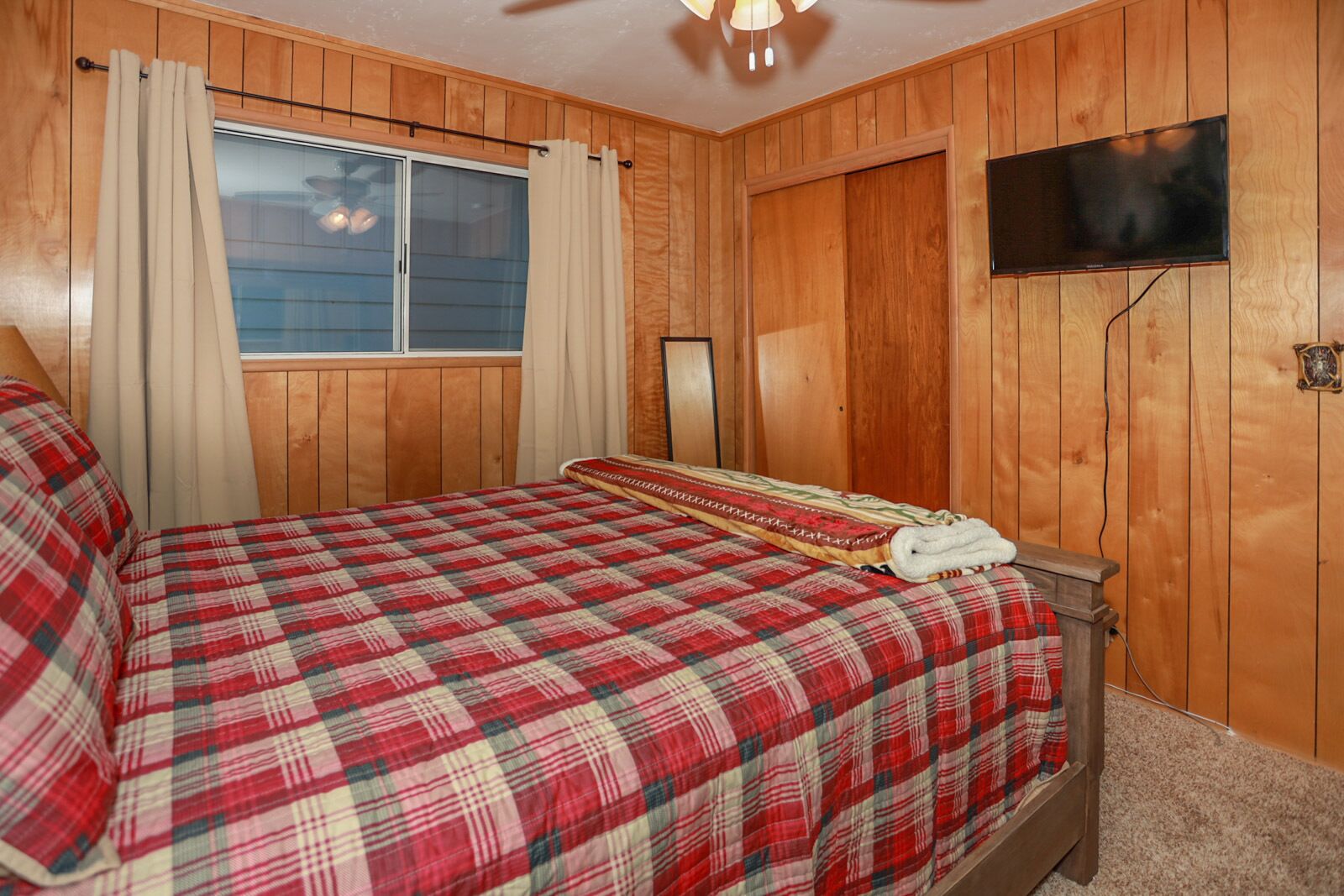 A bedroom with a wooden paneled wall, a bed with a red and white checkered bedspread, a television mounted on the wall, and a window with curtains.