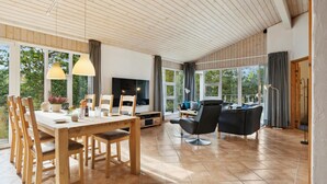 A spacious living area with a wooden dining table, chairs, a sofa, and a TV. Large windows provide natural light and views of greenery. - Lovely home in Fjerritslev with WiFi (Fjerritslev)