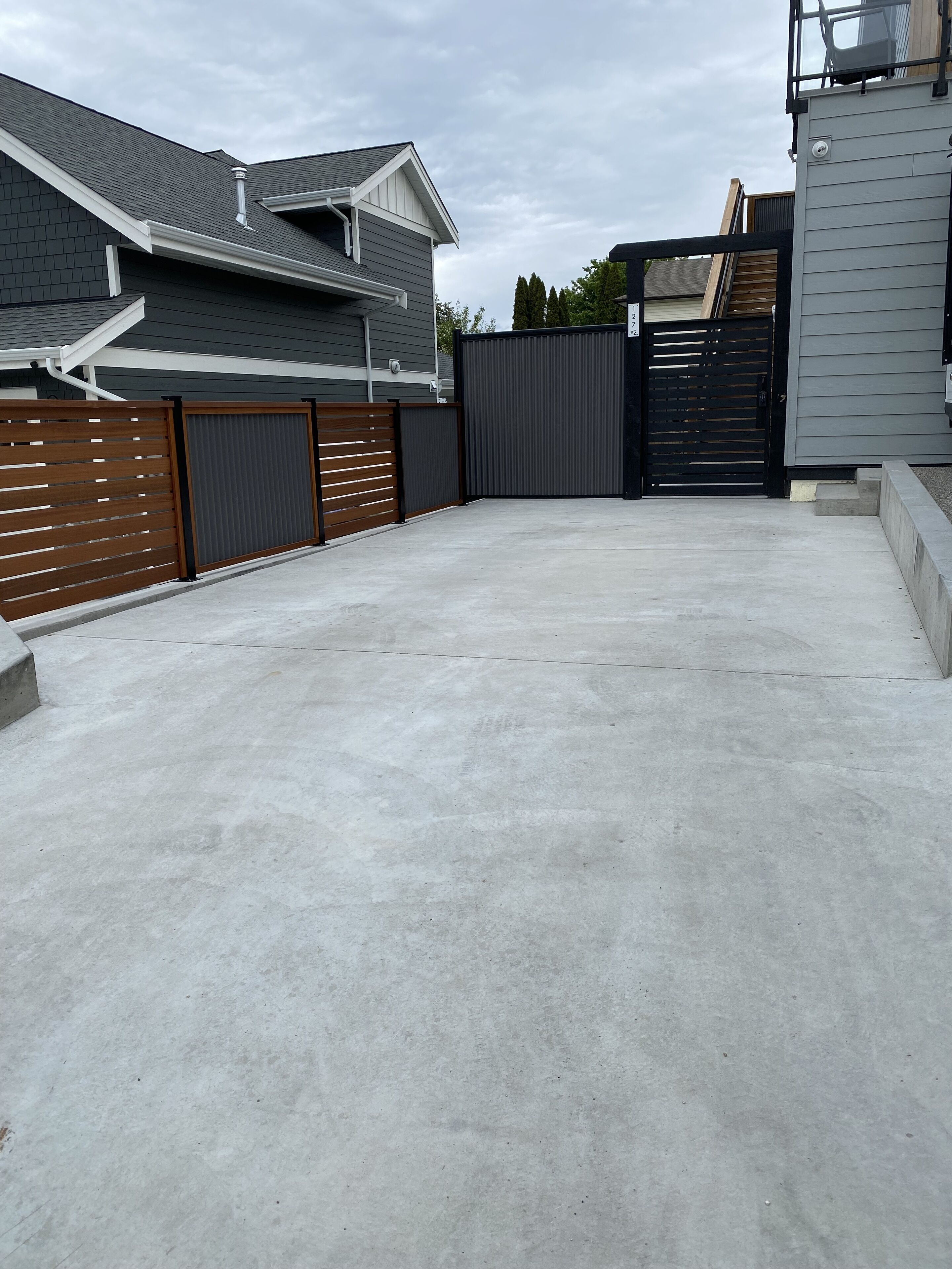 A concrete driveway leading to a gated area with a grey house and a wooden fence.