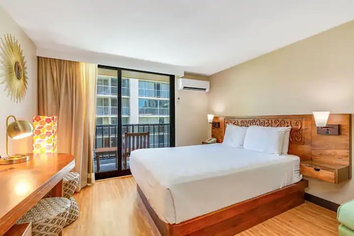 A hotel room with a bed, a desk, a chair, a nightstand, a lamp, and a view of the outside balcony.