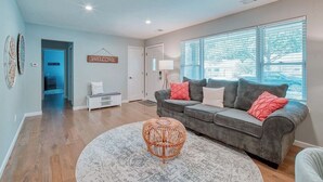 A modern living room with a grey sofa, a round wicker ottoman, and a large window with blinds.