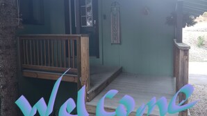 Un banc en bois se trouve devant un bâtiment dont la porte est peinte en vert et sur lequel est affiché un panneau indiquant « BIENVENUE ».