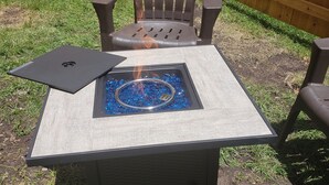 Une table carrée pour l’extérieur, dotée d’un foyer intégré, entourée d’une clôture en bois et d’une zone herbeuse.