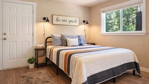 A bedroom with a bed, a bedside table, a wall-mounted lamp, a framed quote, a window with a view of trees, and a door.