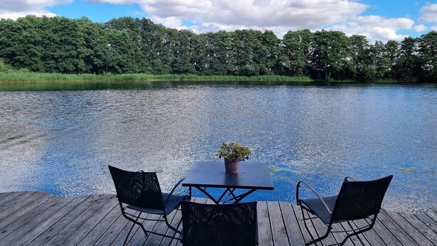 Een houten terras met twee stoelen en een tafel, met uitzicht op een kalm meer met een bebost oever.