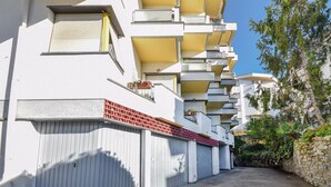 A multi-story residential building with balconies, a gated entrance, and a stone wall on the right.