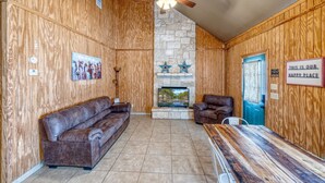 A cozy room with wooden walls, a stone fireplace, a ceiling fan, a brown leather sofa, a brown leather armchair, and a wooden dining table.