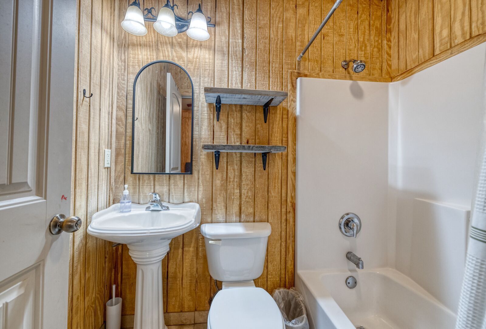 A bathroom with a wooden wall, a pedestal sink, a toilet, a bathtub, and a mirror with lights above.