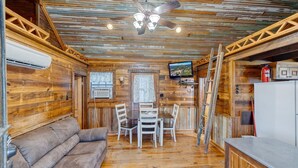 A rustic cabin interior with a wooden ceiling, a ceiling fan, a TV mounted on the wall, a dining table with chairs, and a refrigerator. - Unique Cabin by the Frio - River Rock Cabin #3 (Concan)
