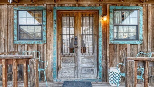 A rustic wooden building with a wooden door, two windows, and a porch with metal chairs and tables. - Unique Cabin by the Frio - River Rock Cabin #3 (Concan)