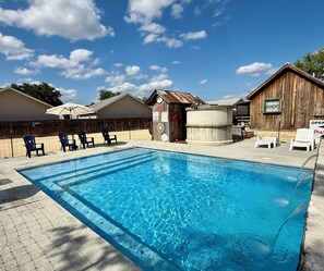 A swimming pool with lounge chairs, a wooden shed, and a fenced area with a wooden building in the background.
