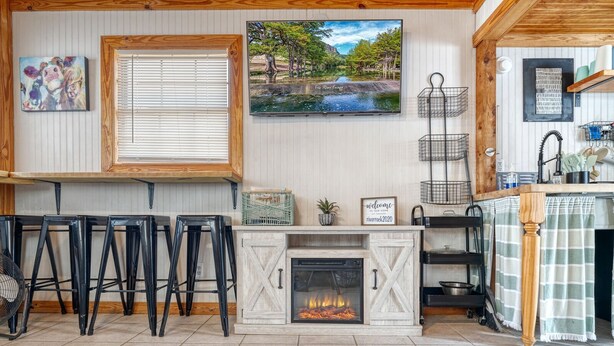 A cozy kitchen with a fireplace, bar stools, a window with blinds, and a painting of a river scene. - Tiny home by the Frio River - River Rock Cabin #4 (Concan)