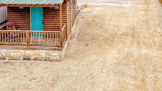 Una cabaña rústica con techo verde, rodeada por un sendero de tierra y vallas de madera, con una colina y cielo despejado al fondo.