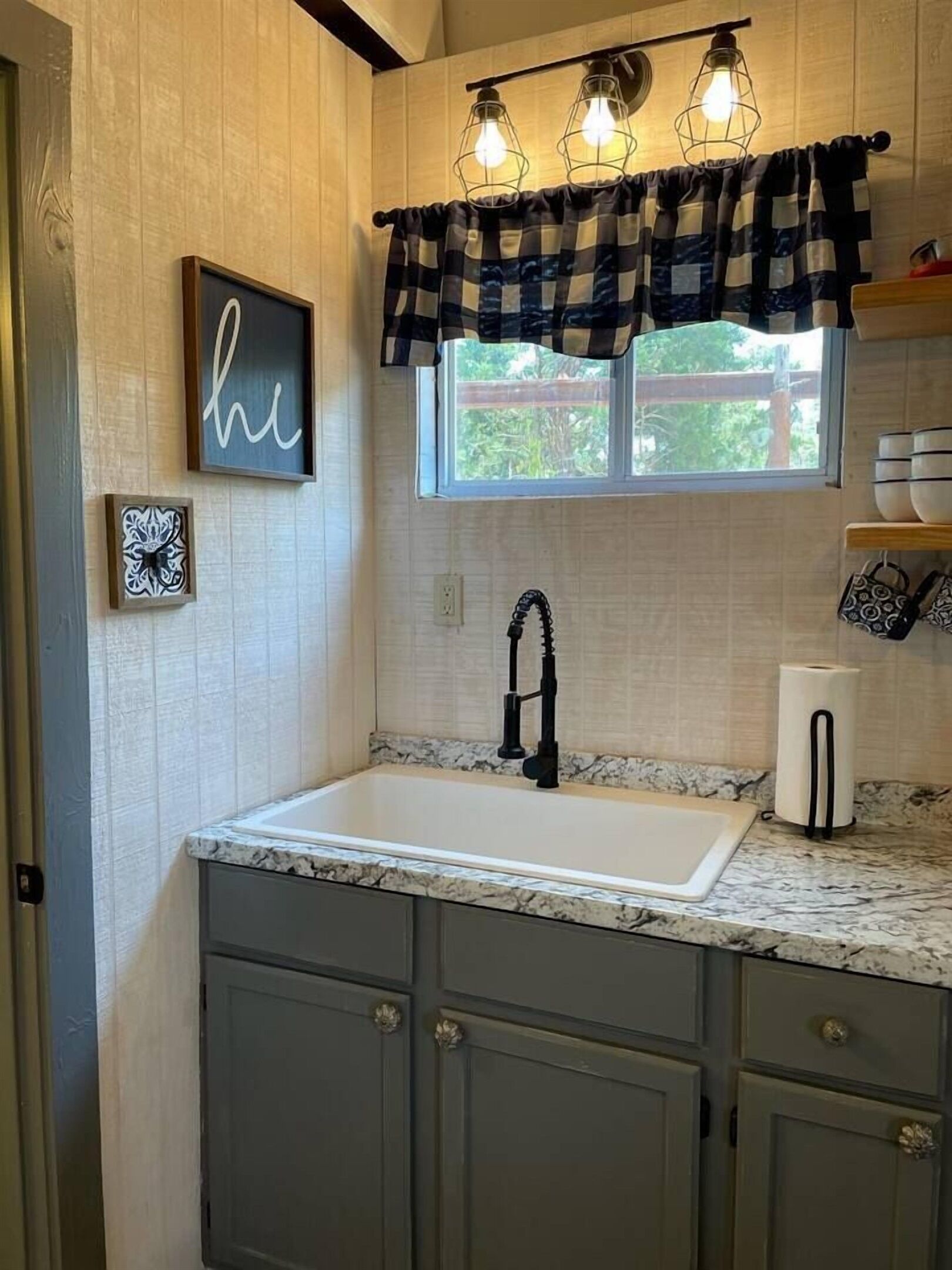 A kitchen with a white sink, dark gray cabinets, a window with a view, and a 'he' sign on the wall.